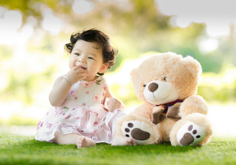 baby sitting on green grass beside bear plush toy at daytime